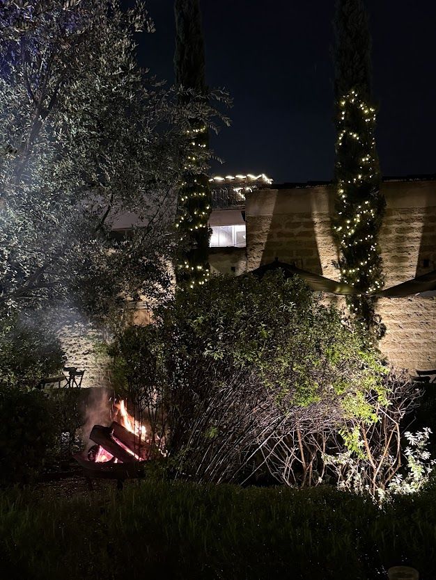 Wood fire in the garden at night, with cypress trees strung with lights against a stone wall